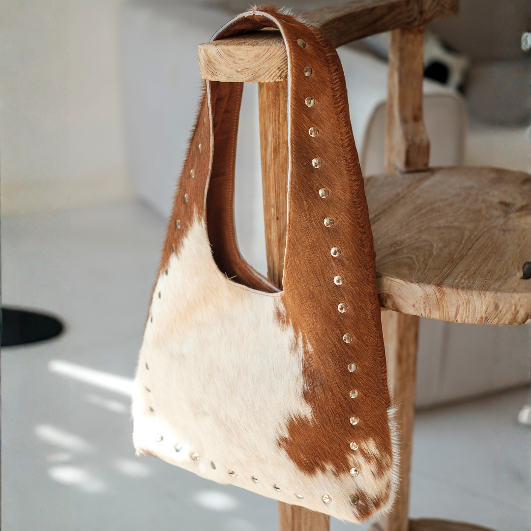 Wrangler Cowhide Bag from House of Hide in brown and cream cowhide leather with stud detailing being displayed hanging on wooden furniture in studio setting