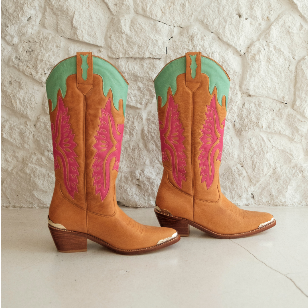 A pair of tan leather cowboy boots with vibrant pink and mint green western embroidery, displayed against a white textured stone wall background.